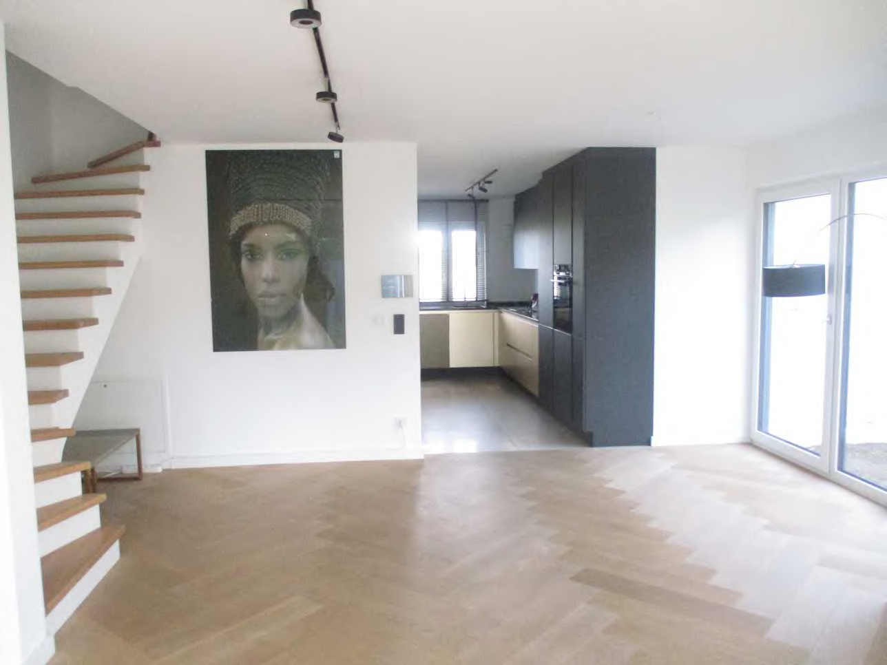 Geräumiger Wohnbereich mit Holzboden, Treppe und Blick in die Küche. An der Wand hängt ein großes Porträt