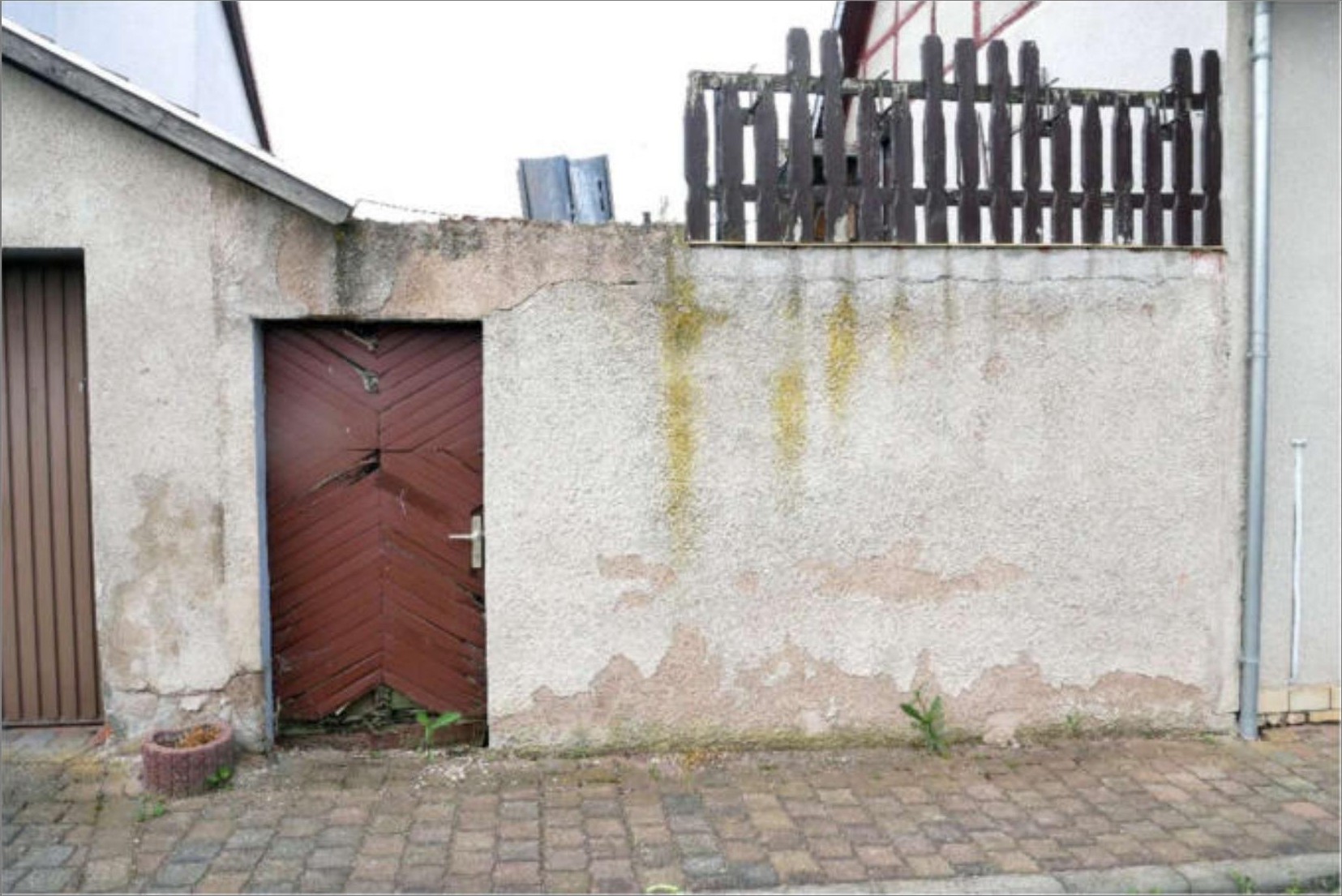 Eingangstür aus rotem Holz in einer verwitterten Betonmauer, gepflasterter Weg davor, kleiner Holzzaun oben