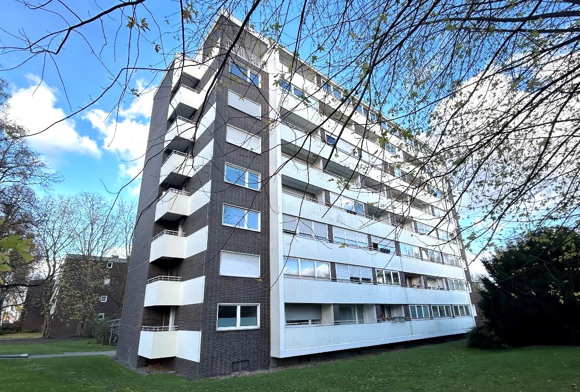 Seitenansicht eines mehrstöckigen Wohnhauses mit Balkonen und Fenstern, umgeben von Grünanlagen unter blauem Himmel