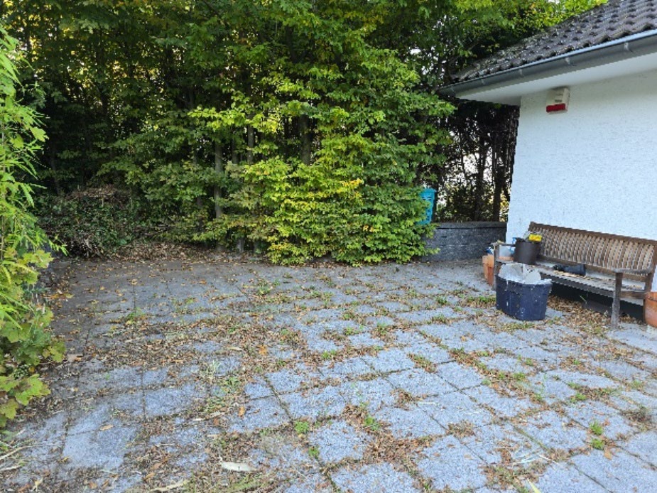 Pflasterter Gartenbereich mit Bäumen und Sträuchern, Holzbank und Mülltonnen neben einem weißen Gebäude mit Ziegeldach