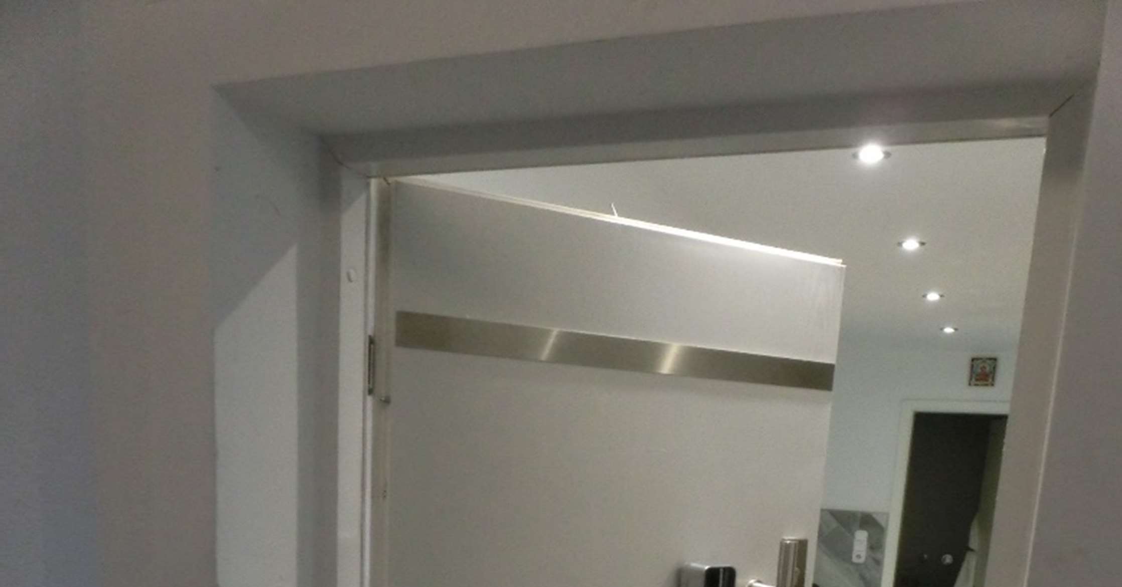 Innenraumansicht durch eine Türöffnung mit weißer Wand, eingelassenen Deckenleuchten und einem metallischen Streifen an der Wand
