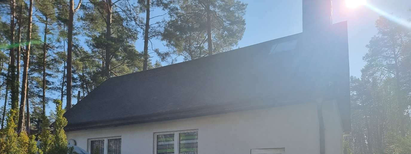Seitenansicht eines weißen Gebäudes mit dunklem Dach, umgeben von Nadelbäumen, bei hellem Tageslicht