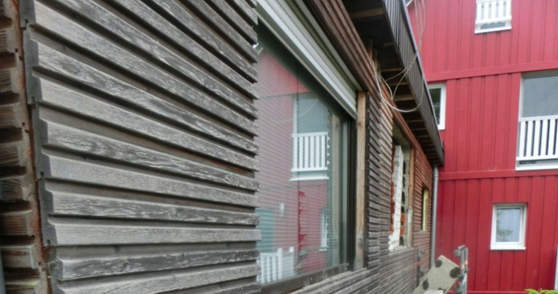 Seitenansicht eines Einfamilienhauses mit verwittertem Holzschindelverkleidung und großem Fenster, daneben ein rotes Gebäude