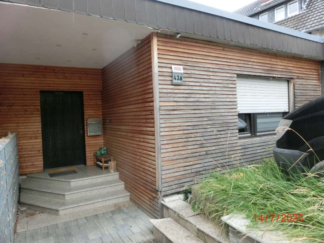 Eingangsbereich eines Einfamilienhauses mit Holzfassade, schwarzer Haustür, Betonstufen und Hausnummer 43a
