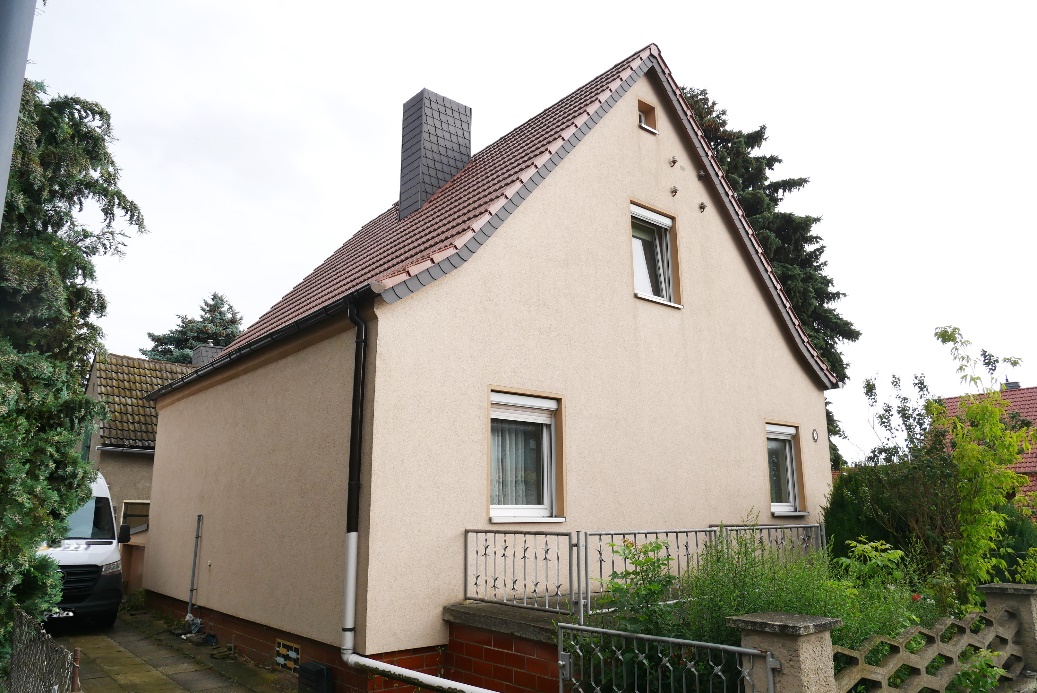 Seitenansicht eines Einfamilienhauses mit Satteldach, Fenstern und Schornstein, umgeben von Grünanlagen und einem Metallzaun