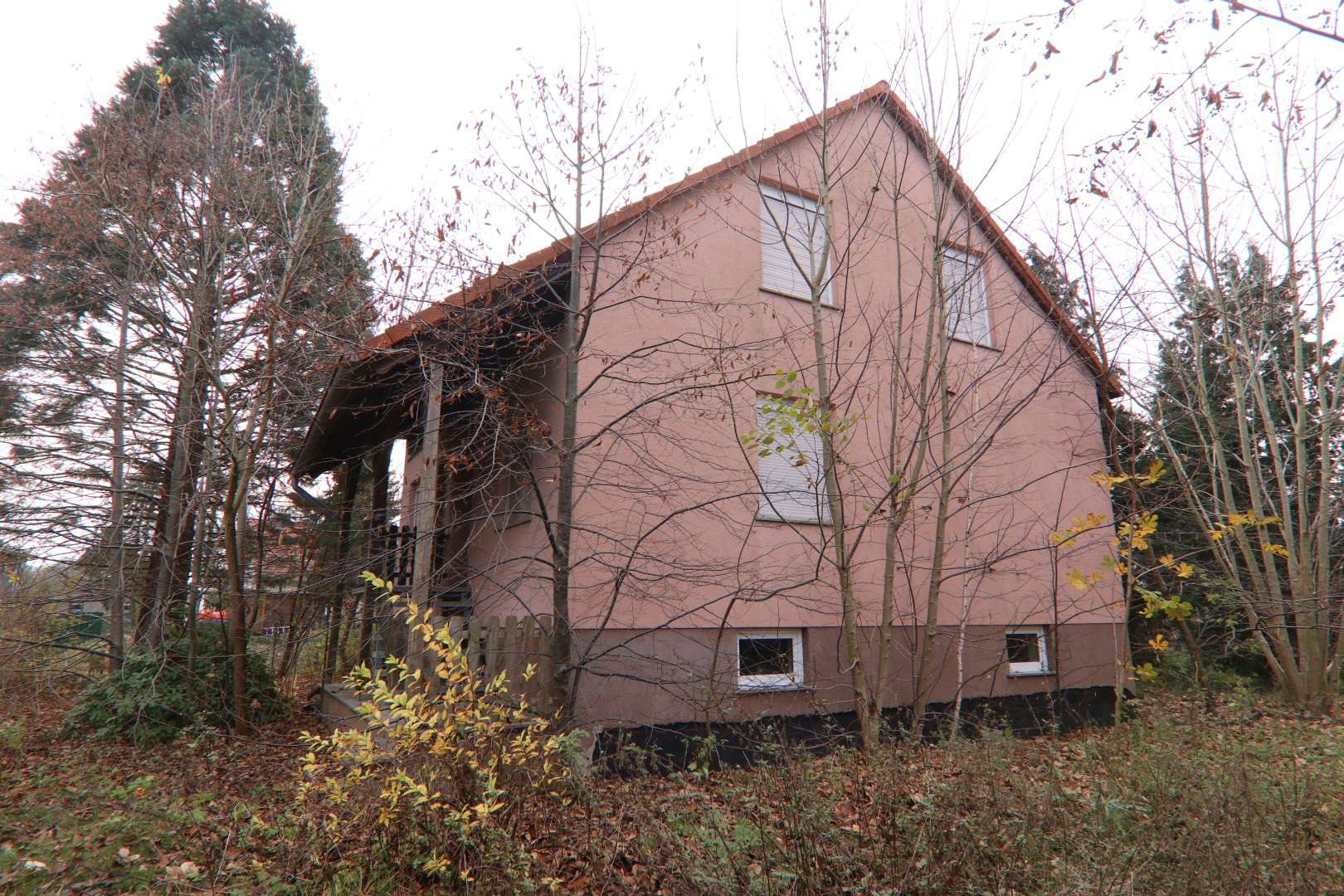 Seitenansicht eines rosa verputzten Gebäudes mit Satteldach, umgeben von kahlen Bäumen und Laub im Herbst. Eingangsbereich mit Holzveranda links