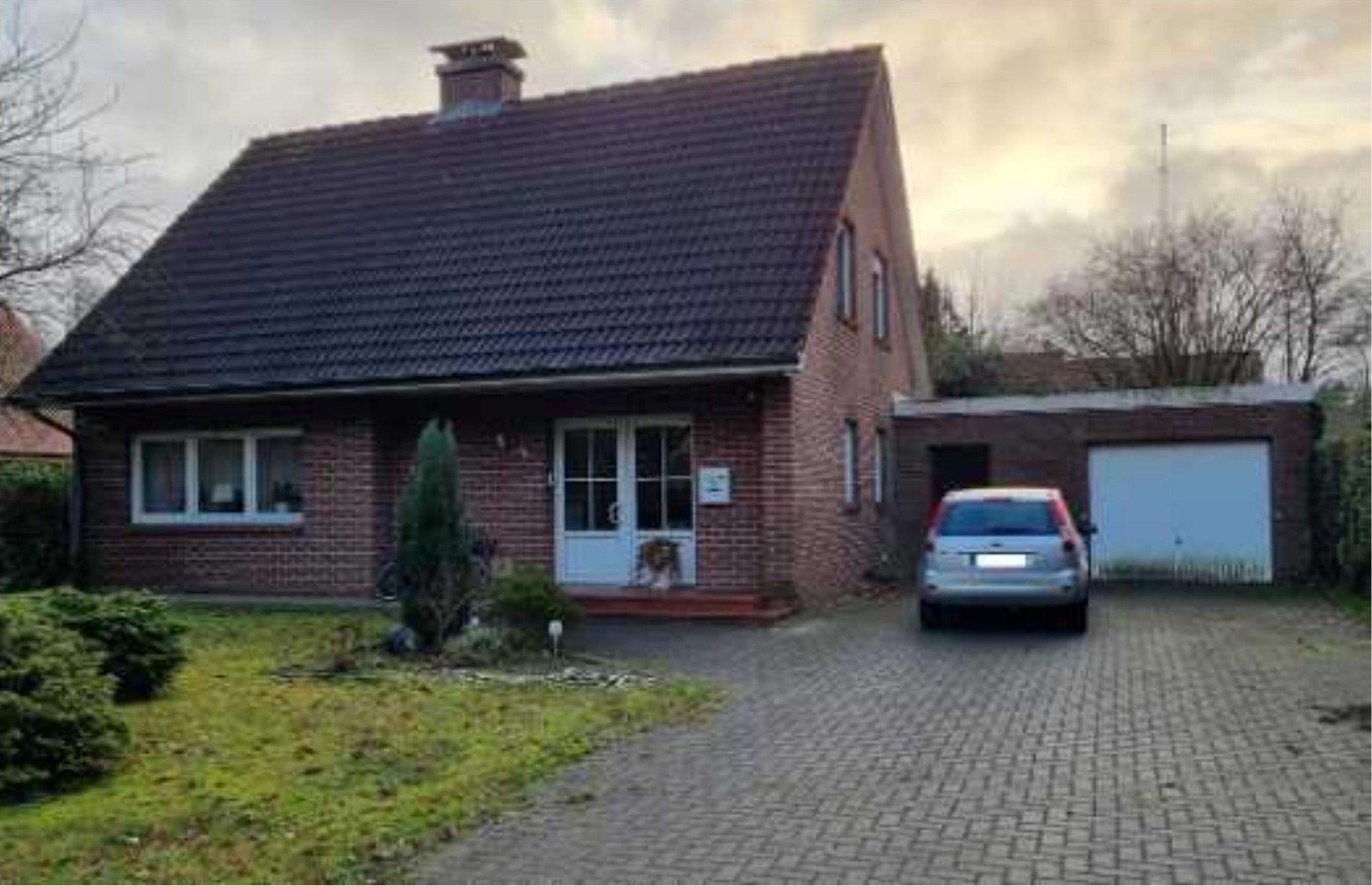 Einfamilienhaus aus Backstein mit Satteldach, Eingangstür, Fenstern und angrenzender Garage. Vor dem Haus befindet sich ein gepflasterter Vorplatz mit einem geparkten Auto und