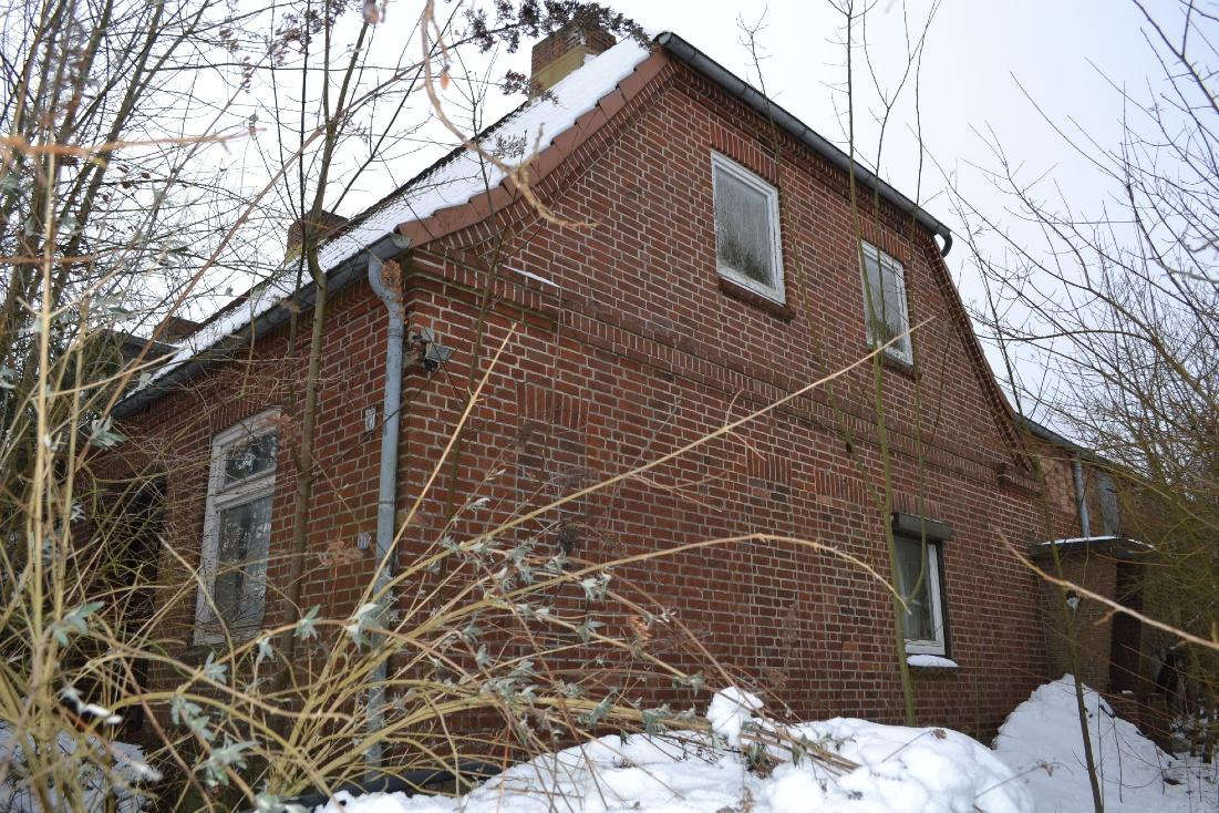 Seitenansicht eines Backsteinhauses mit Schnee auf dem Dach und im Garten, umgeben von kahlen Sträuchern