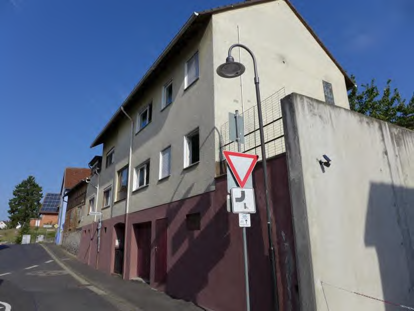 Seitenansicht eines zweistöckigen Wohnhauses mit grauer Fassade und rotem Sockelbereich, Straßenlaterne und Verkehrszeichen am Rand einer Straße in Bad Nauheim