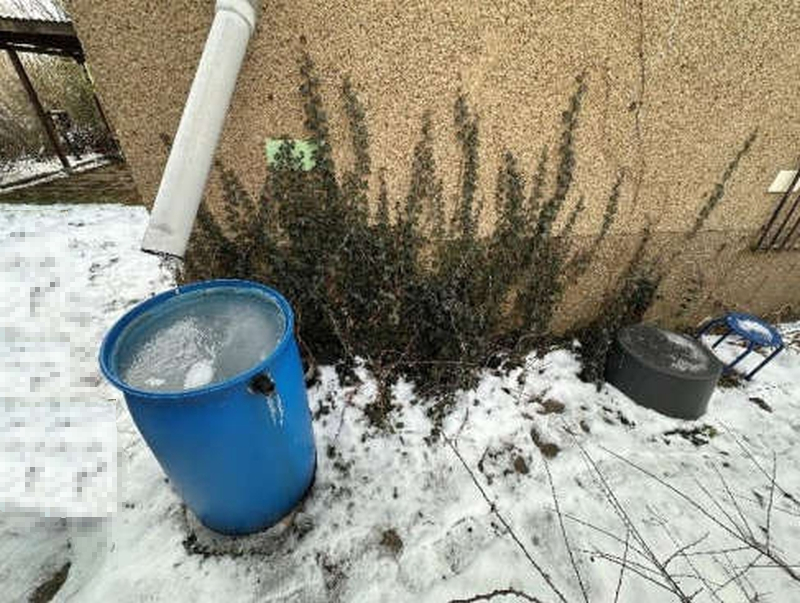 Schneebedeckter Gartenbereich mit blauem Eimer, der mit Eis gefüllt ist, und einem Regenrohr an einer verputzten Wand