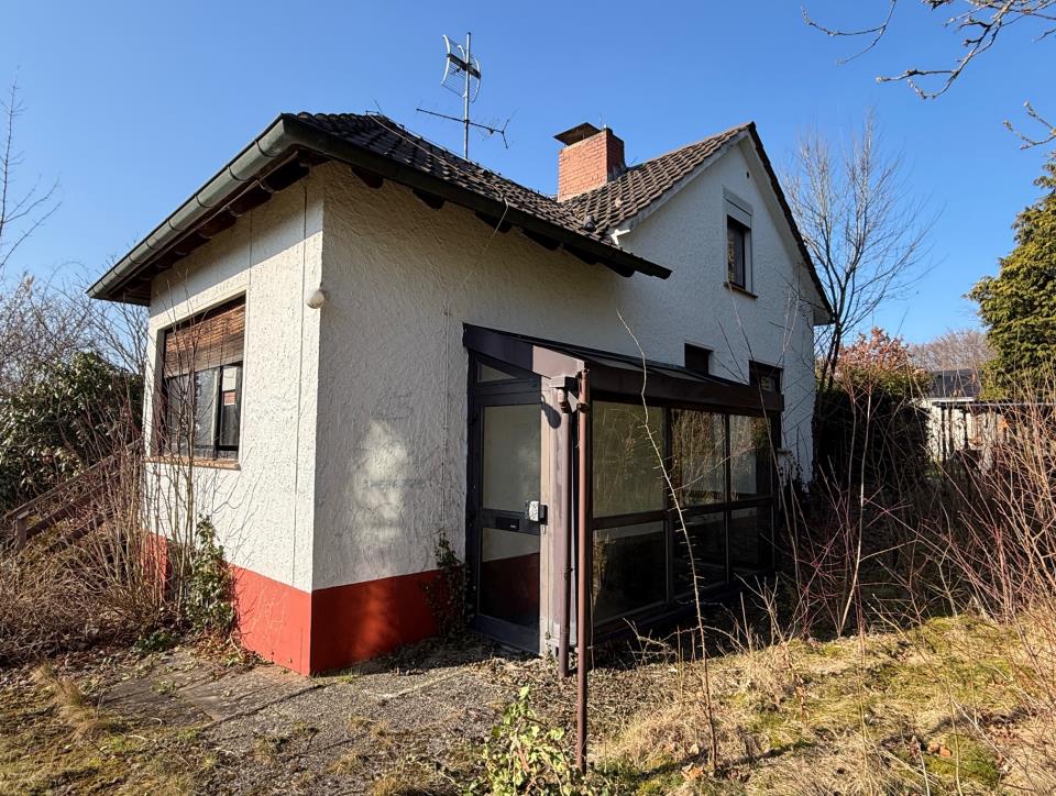 Einfamilienhaus mit weißer Fassade und rotem Sockel, Eingang mit Glasverglasung, Dach mit Schornstein und Antenne, Garten im Vordergrund