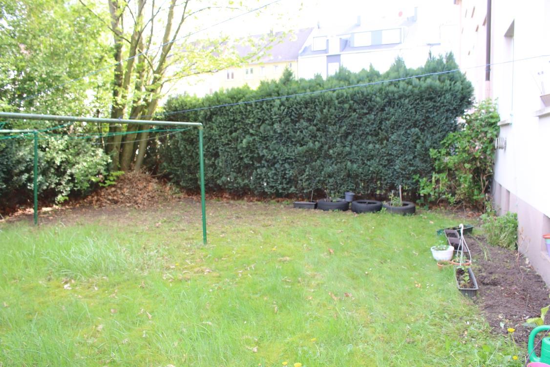 Grüner Gartenbereich mit Rasen, Hecken, Bäumen und Gartenutensilien wie Reifen und Pflanzkübeln an der Seite eines Gebäudes
