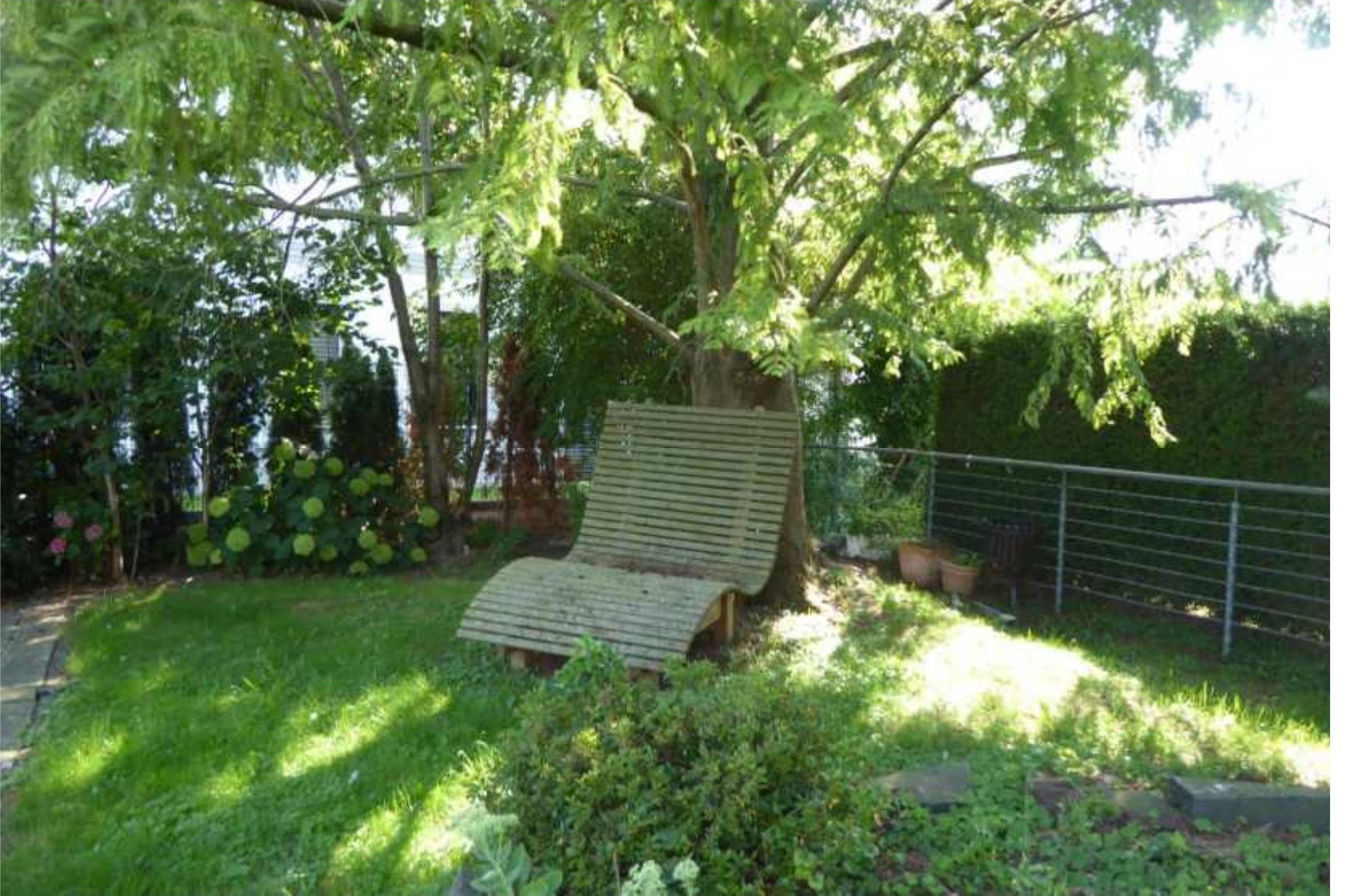 Gartenbereich mit Gras, Bäumen, Sträuchern und einer Holzbank unter einem Baum. Metallgeländer im Hintergrund