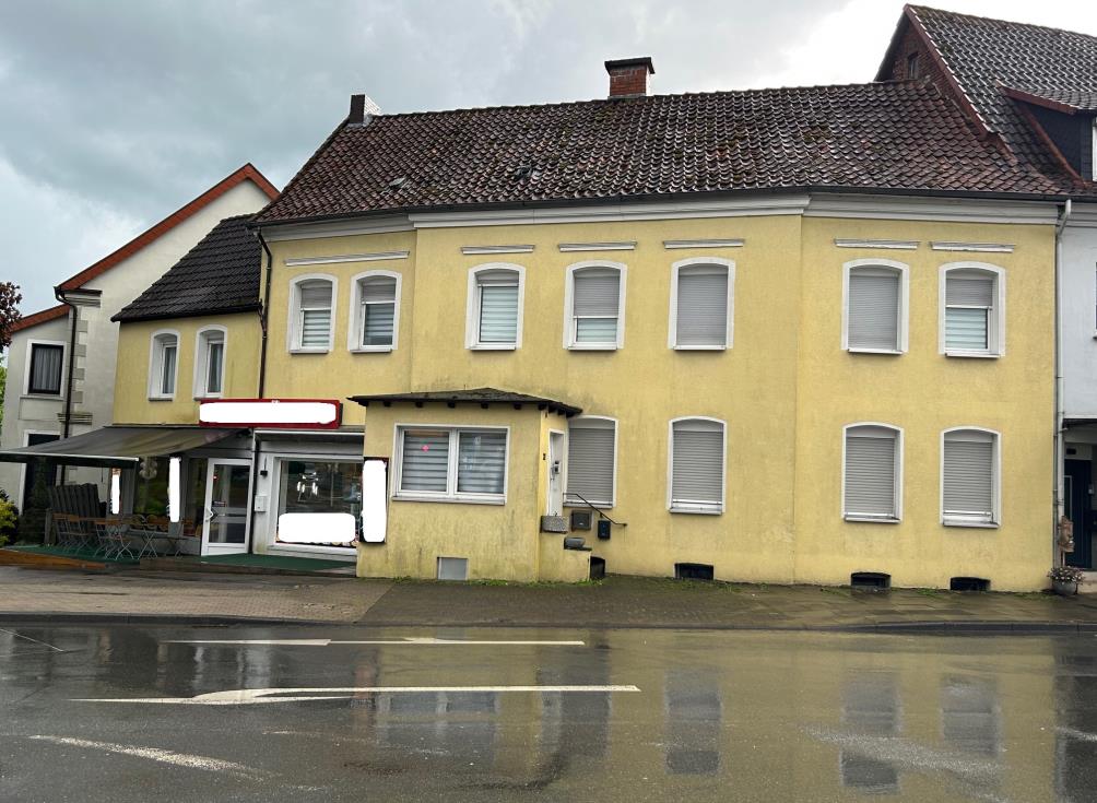 Gelbes Wohn- und Geschäftshaus mit Satteldach und mehreren Fenstern, Ansicht von vorne, nasser Straßenbelag im Vordergrund