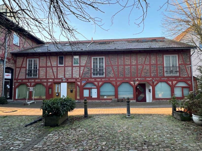 Vorderansicht eines zweistöckigen Fachwerkhauses mit roten Holzbalken und Ziegeldach, Eingangstüren und Fenstern, gepflastertem Vorplatz und Pflanzenkästen