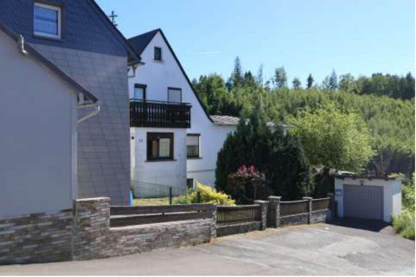 Seitenansicht eines Einfamilienhauses mit Balkon, Steinmauer und Gartenbereich, im Hintergrund Bäume und ein Garagenanbau