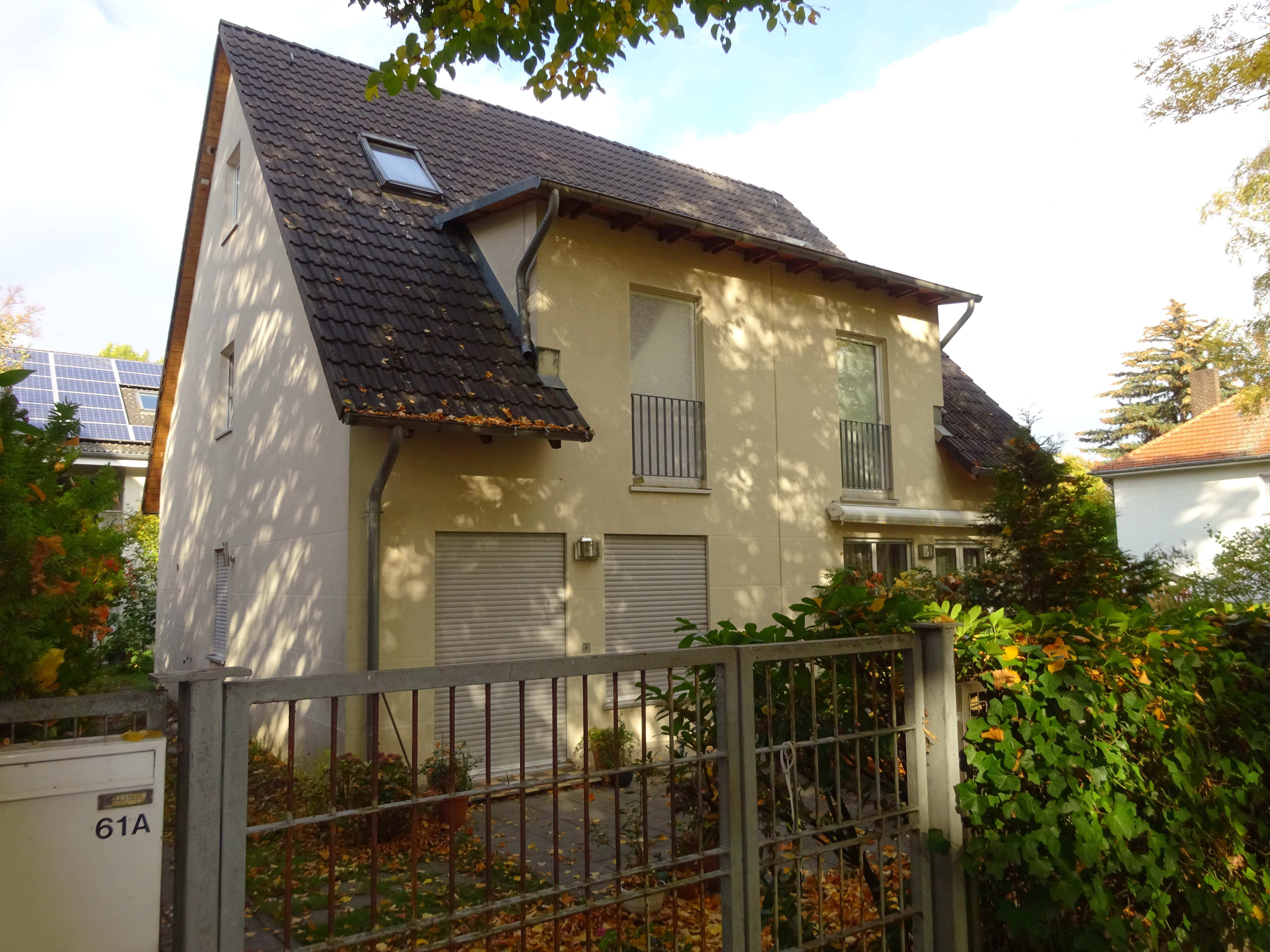 Vorderansicht einer Doppelhaushälfte mit gelber Fassade, dunklem Satteldach und Garagentoren, umgeben von Hecke und Metallzaun, Adresse Oehlertring 61