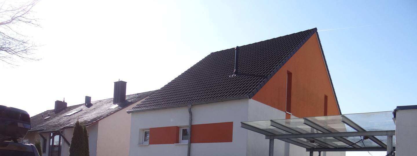 Seitenansicht eines Einfamilienhauses mit schwarzem Ziegeldach, weißer und orangefarbener Fassade sowie einem Glasdachüberstand