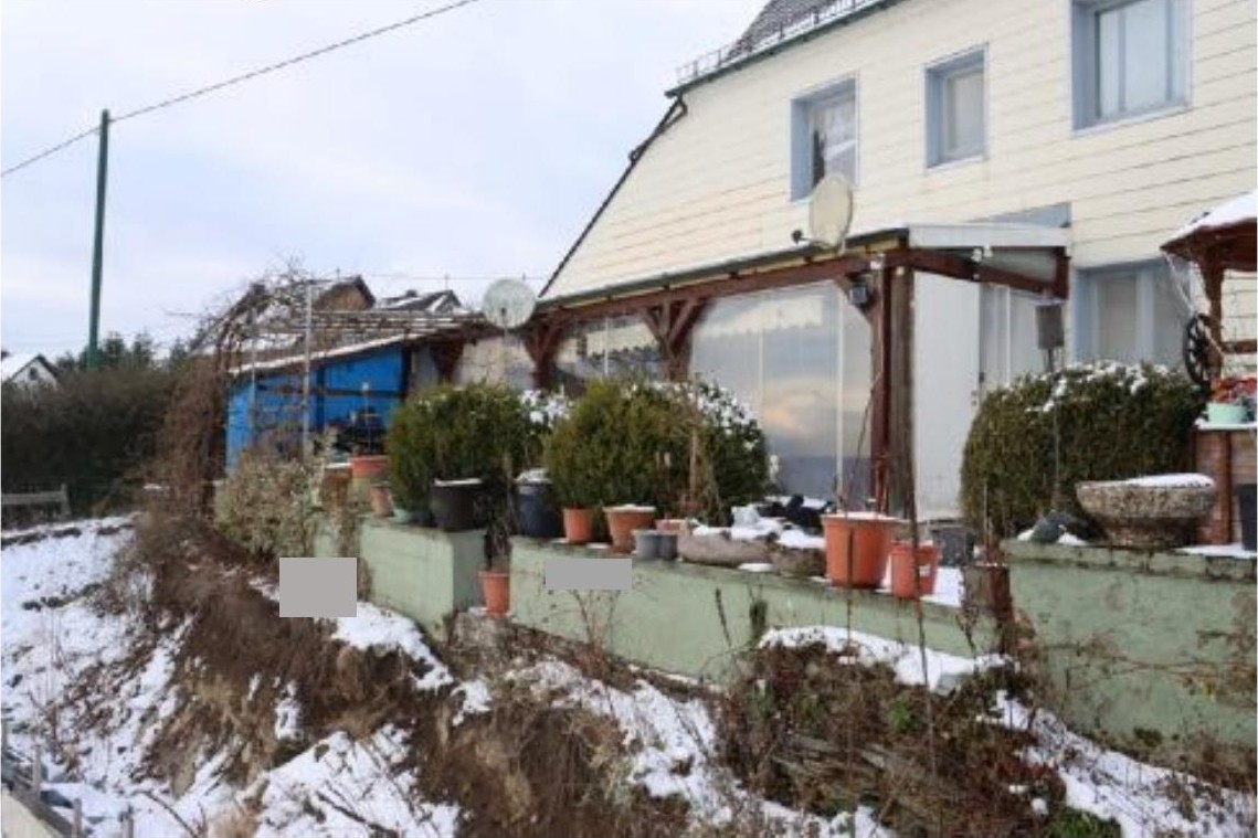 Gartenbereich mit Pflanzen in Töpfen, niedriger Mauer und Schneebedeckung vor einem Haus mit Terrasse in Leienkaul