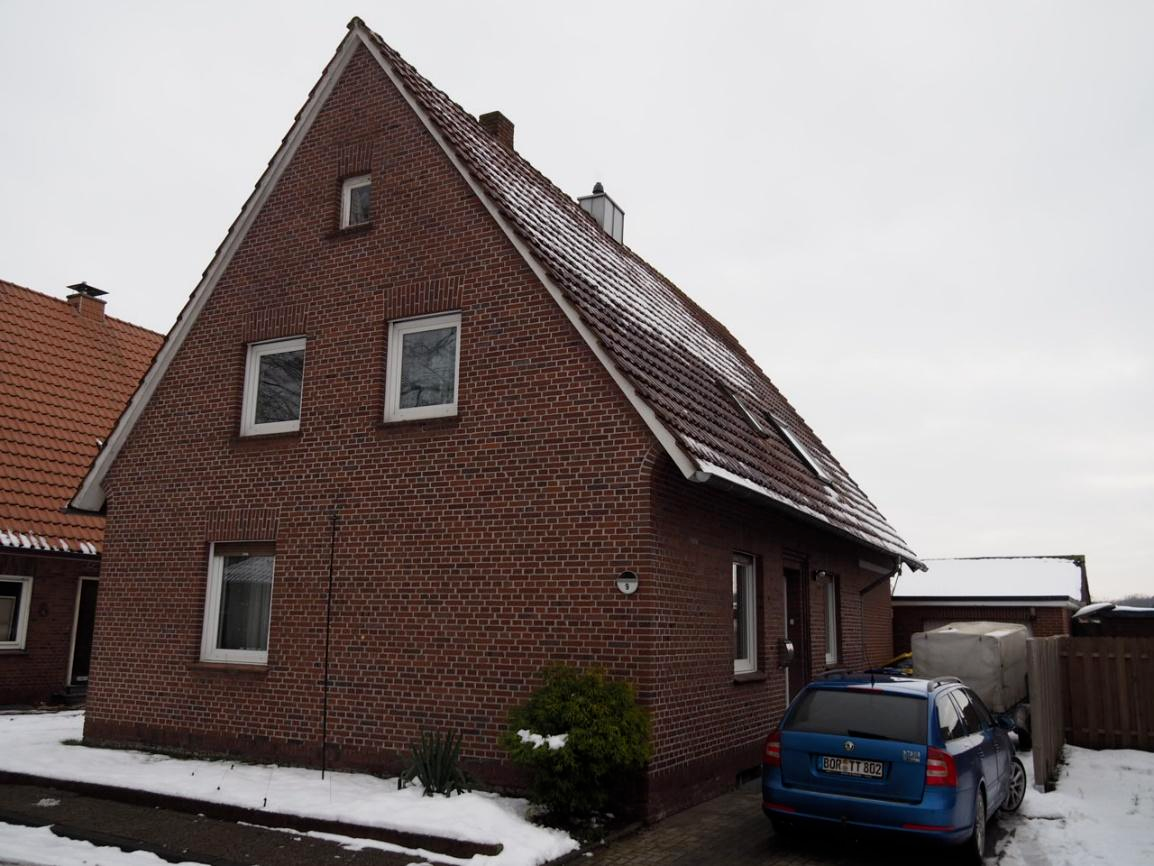 Einfamilienhaus aus rotem Backstein mit Satteldach, mehreren Fenstern und Eingangstür; vorne geparkter blauer Kombi, Schnee auf Boden und Dach