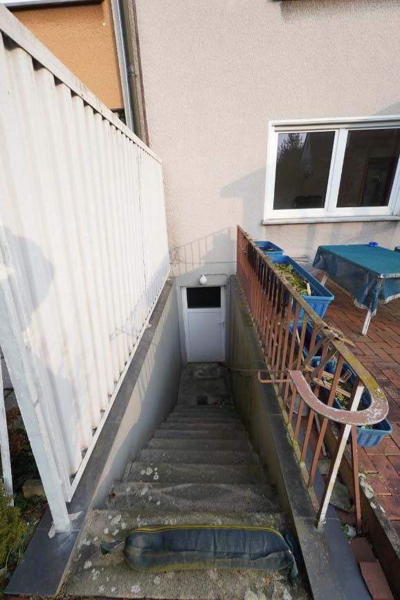 Außenansicht einer Betontreppe mit Geländer, die zu einer weißen Tür im Erdgeschoss führt, neben einem gepflasterten Terrassenbereich