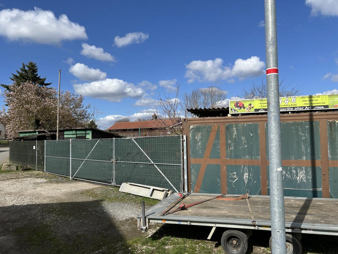 Seitenansicht eines Grundstücks mit Metallzaun, Holzcontainer und Anhänger im Vordergrund, dahinter Gebäude und Bäume unter blauem Himmel