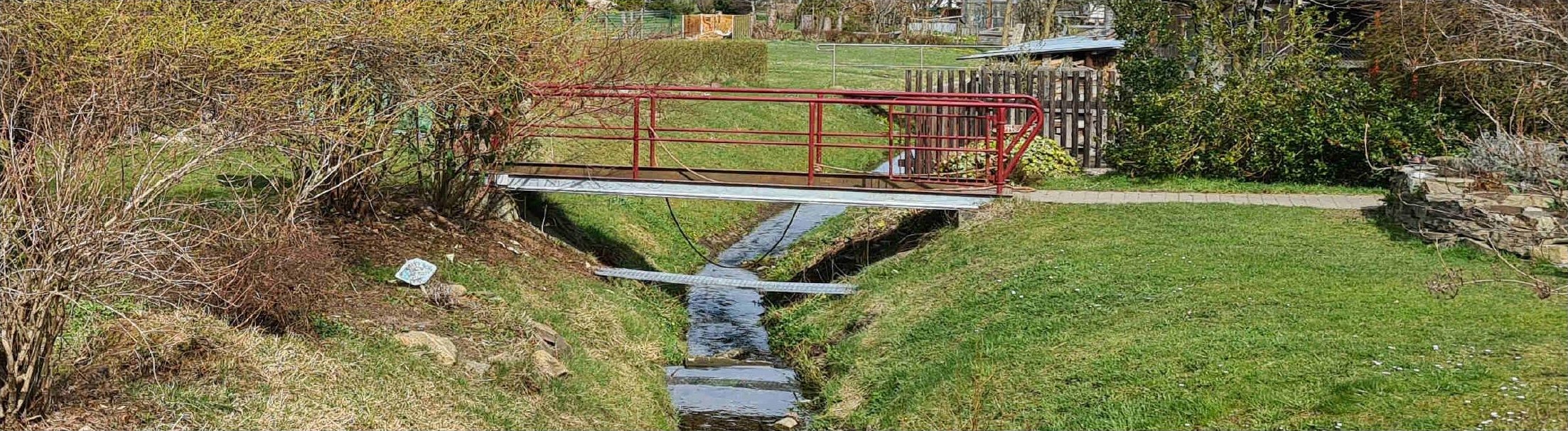 Gartenbereich mit rotem Metallbrückchen über einem kleinen Bach, grünen Wiesen und Sträuchern im Vordergrund