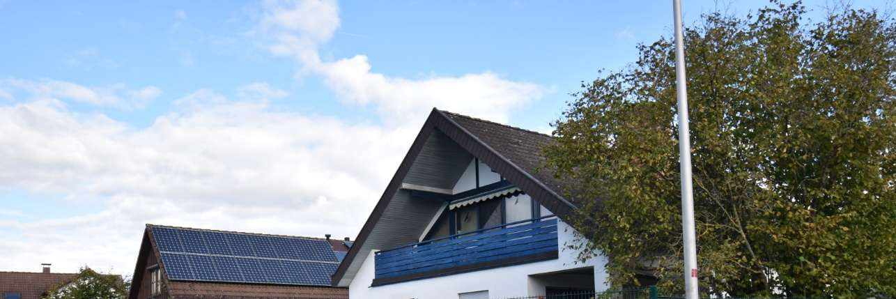 Seitenansicht eines Gebäudes mit Dachgaube, blauem Balkon und Solaranlage auf dem Dach unter bewölktem Himmel