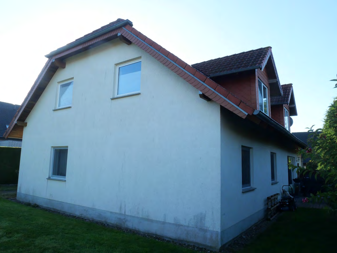 Seitenansicht eines zweistöckigen Wohnhauses mit hellen Wänden, rotem Ziegeldach und Fenstern; Grünfläche im Vordergrund