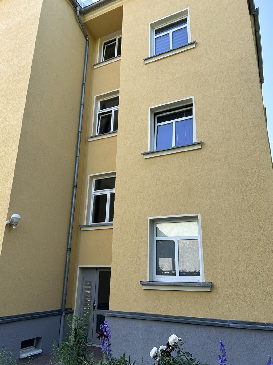 Seitenansicht eines gelb gestrichenen Mehrfamilienhauses mit weißen Fenstern und grauem Sockelbereich, Eingangstür sichtbar, vorne Pflanzen