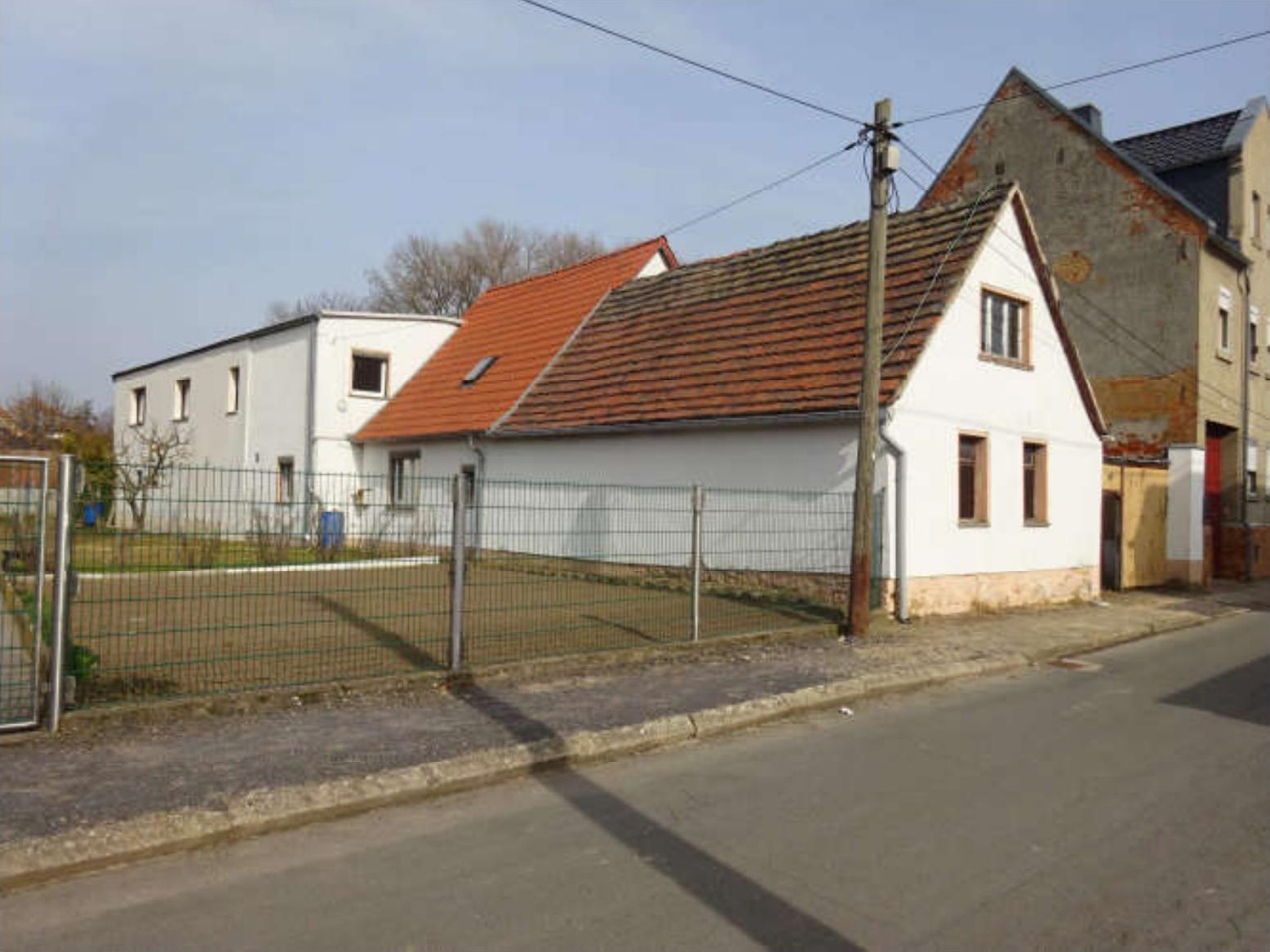 Seitenansicht eines weißen Gebäudes mit rotem Ziegeldach in Teuchern OT Nessa, Kapellenende 69, hinter einem Metallzaun und neben einer Straße