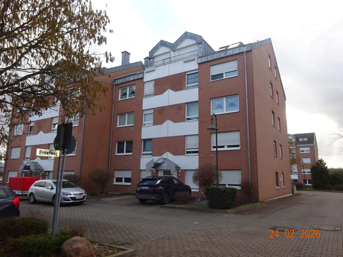 Seitenansicht eines mehrstöckigen Wohngebäudes aus rotem Backstein mit weißen Fassadenelementen und Fenstern, geparkte Autos im Vordergrund, Straßenschild Birkenweg sichtbar