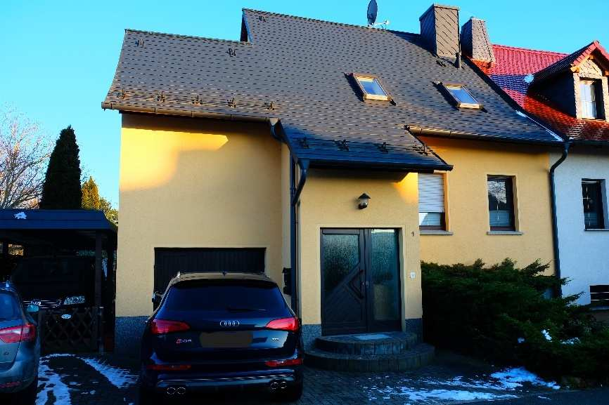 Vorderansicht einer Doppelhaushälfte mit gelber Fassade, dunklem Dach und Eingangstür; vor dem Haus geparkte Autos und Schneereste auf dem Kopfsteinpflaster