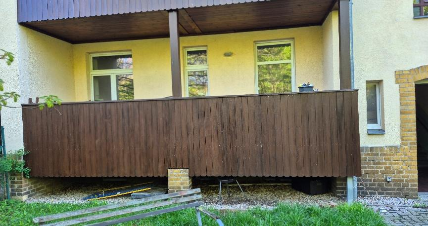 Holzbalkon an der Fassade eines gelb gestrichenen Gebäudes mit Fenstern und Säulenstützen, im Hintergrund Grünfläche