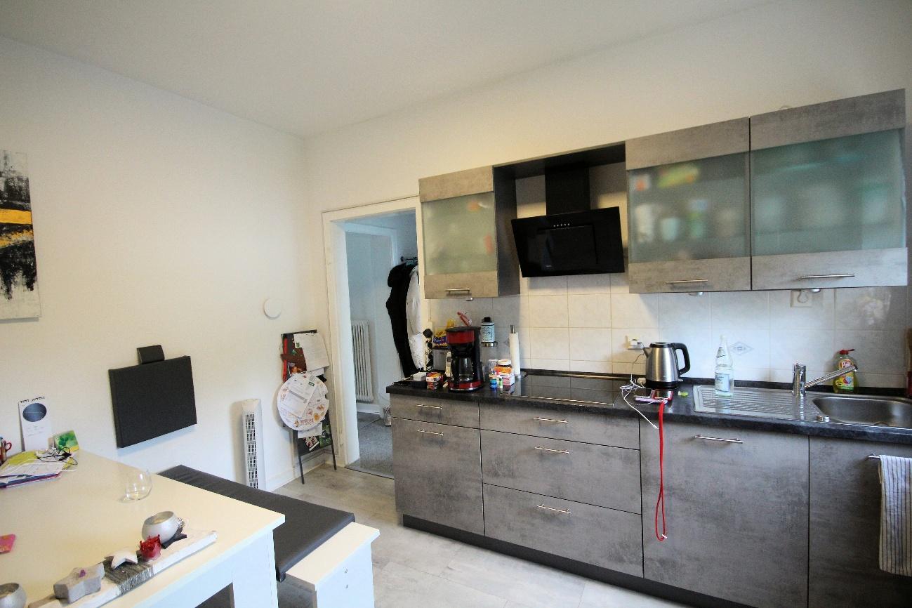 Innenraum eines Wohnbereichs mit integrierter Küche, grauen Schränken, schwarzer Arbeitsplatte und einem Essbereich mit weißem Tisch und TV