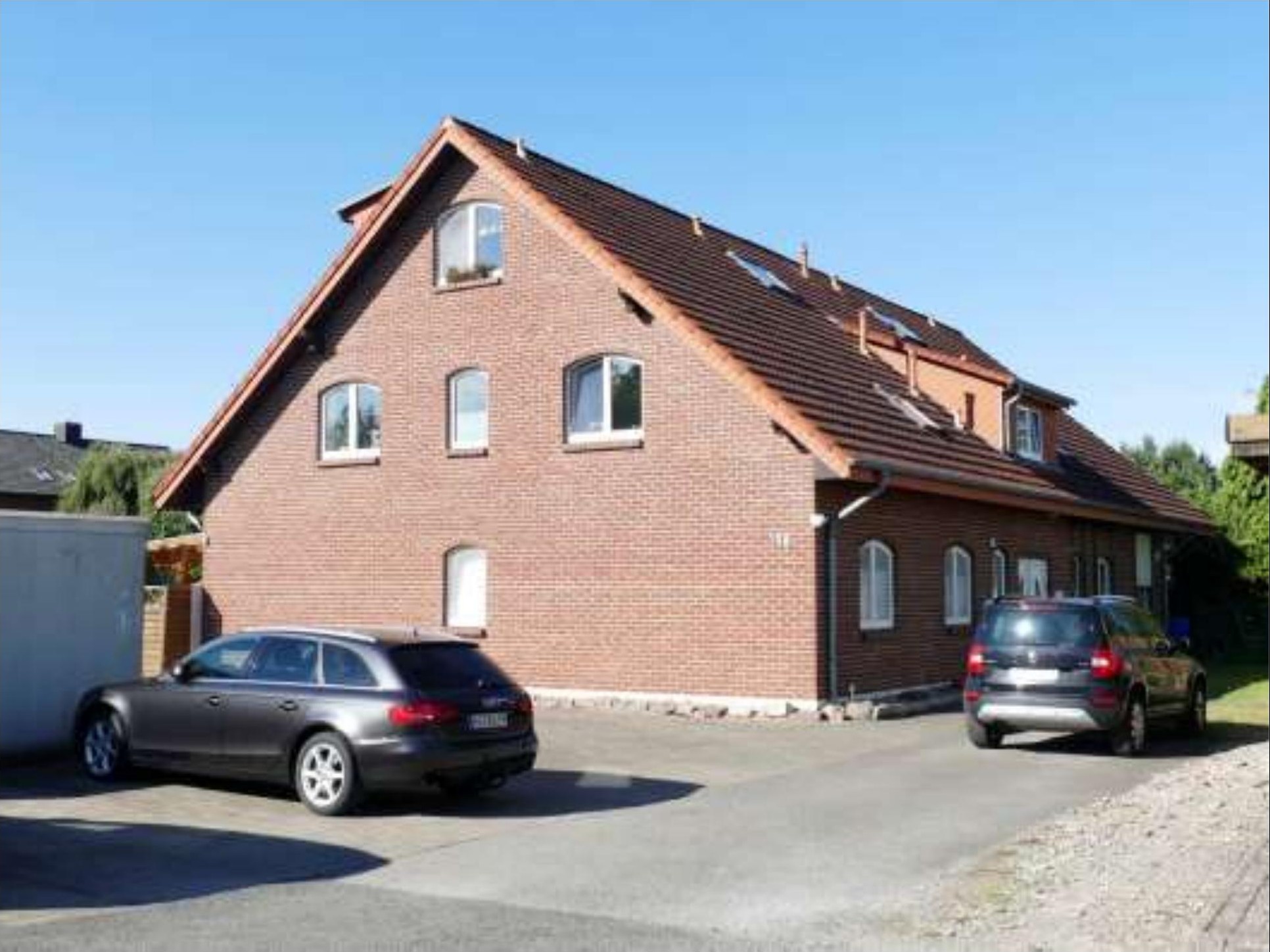 Seitenansicht eines zweistöckigen Backsteinhauses mit Satteldach, mehreren Fenstern und zwei geparkten Autos vor dem Gebäude