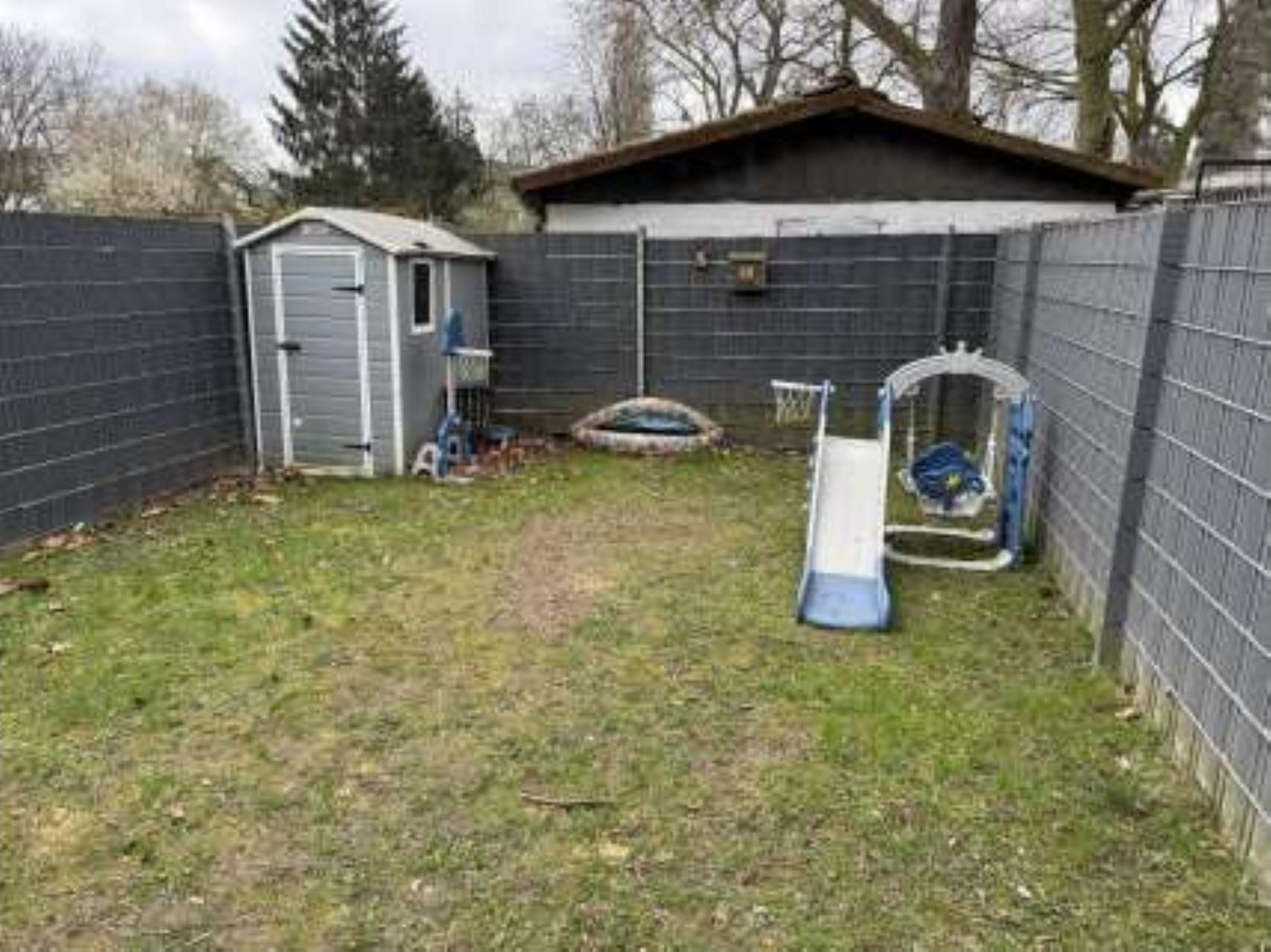 Hinterhof mit Rasenfläche, Kinderspielgerät, Gartenhaus und grauer Umzäunung