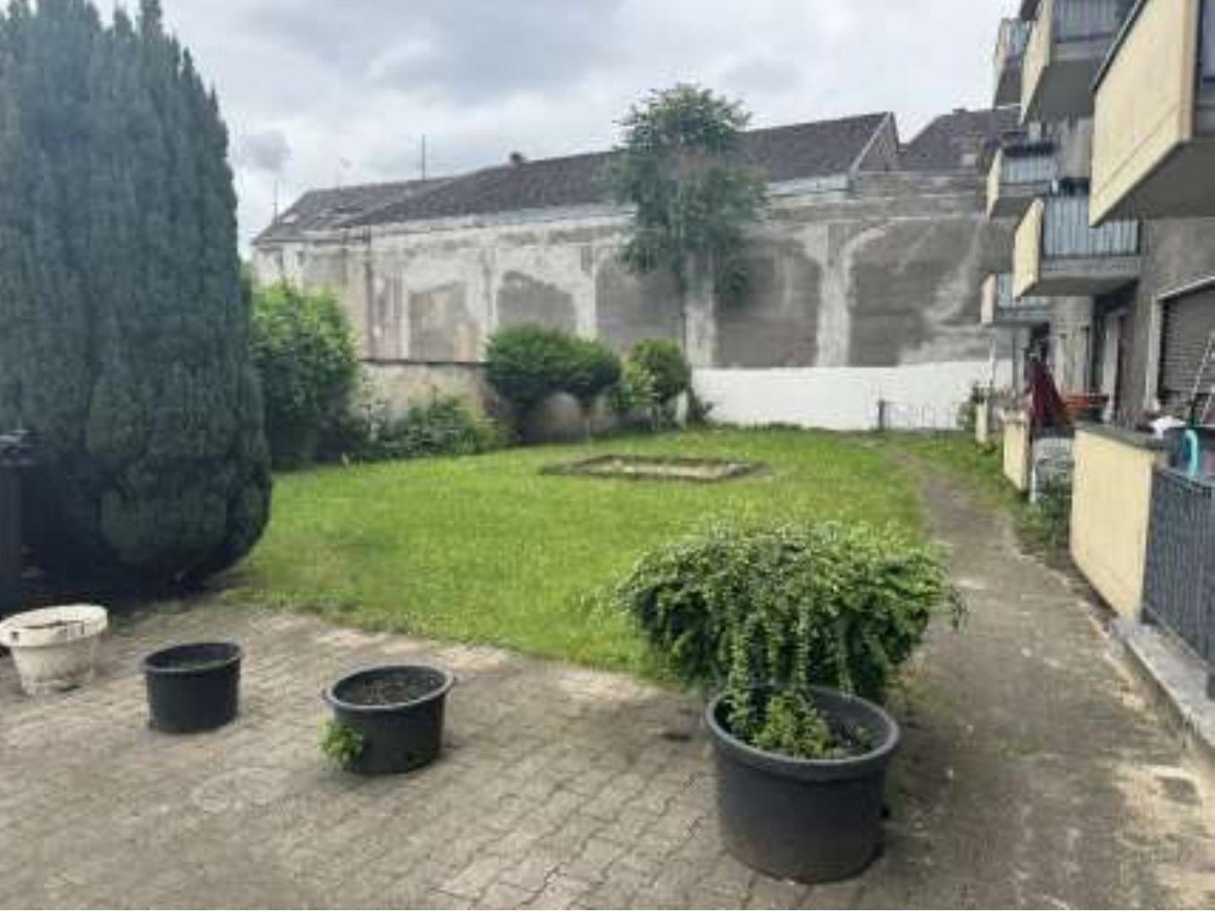 Gartenbereich mit Rasenfläche, Pflanzkübeln und einem gepflasterten Weg neben einer Wohnanlage in Duisburg