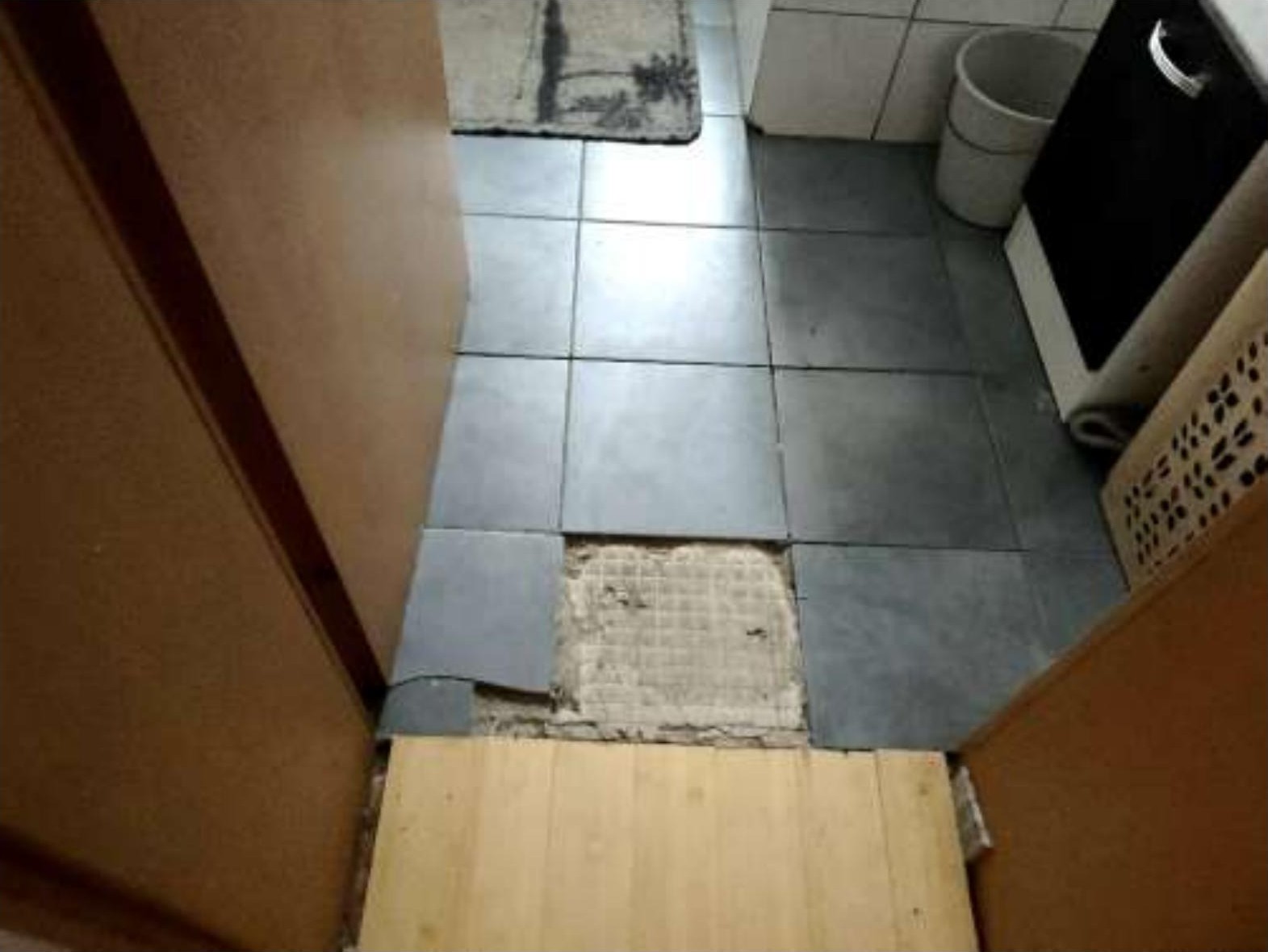 Badezimmerboden mit grauen Fliesen, einer beschädigten Fliese, einem Teppich und einem Mülleimer. Teil einer Waschmaschine oder Trockner sichtbar