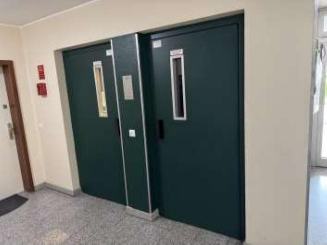 Innenansicht eines Flurs mit zwei dunkelgrünen Türen, Fliesenboden und Wandmontage von Feueralarmgeräten in einer Eigentumswohnung in Duisburg