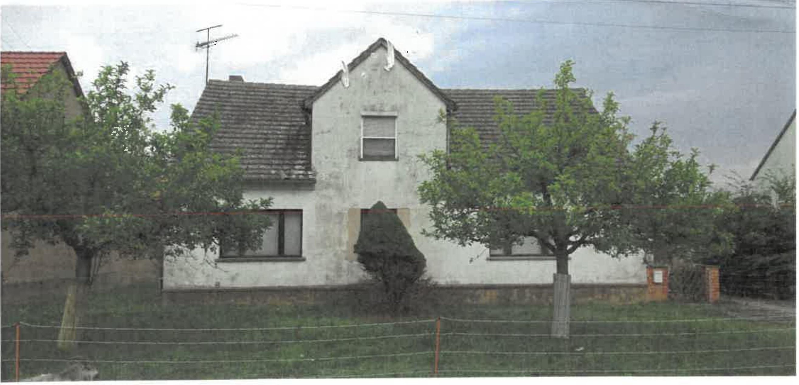 Einfamilienhaus mit weißer Fassade, Satteldach und Vorgarten, umgeben von Bäumen und einem Drahtzaun