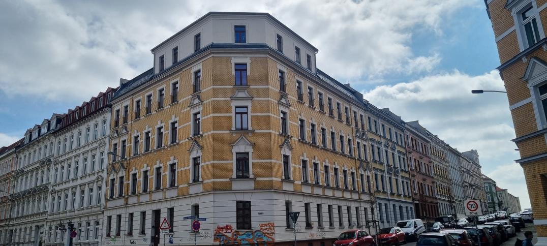 Gebäudeecke einer mehrstöckigen Wohnhausfassade in gelb-weißer Fassade mit Fenstern und Dachgauben, Straßenansicht mit geparkten Autos