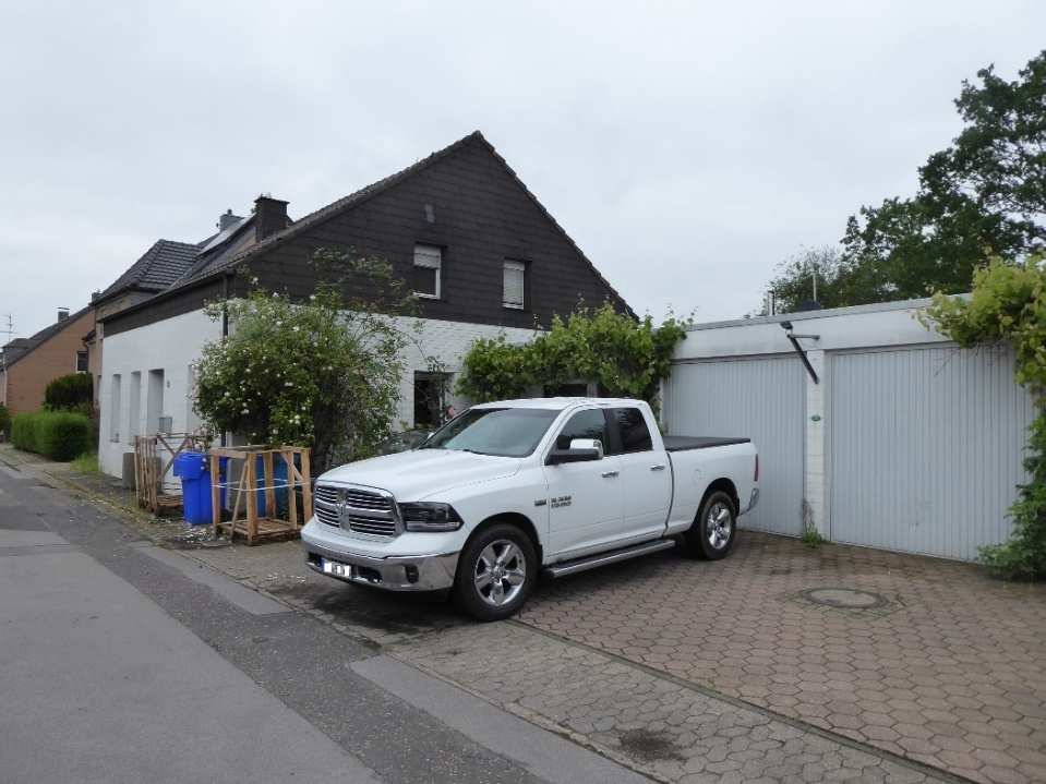 Einfamilienhaus mit dunklem Satteldach und weißer Fassade, vorne geparkter weißer Pickup, Garagenanbau rechts, gepflasterter Vorplatz