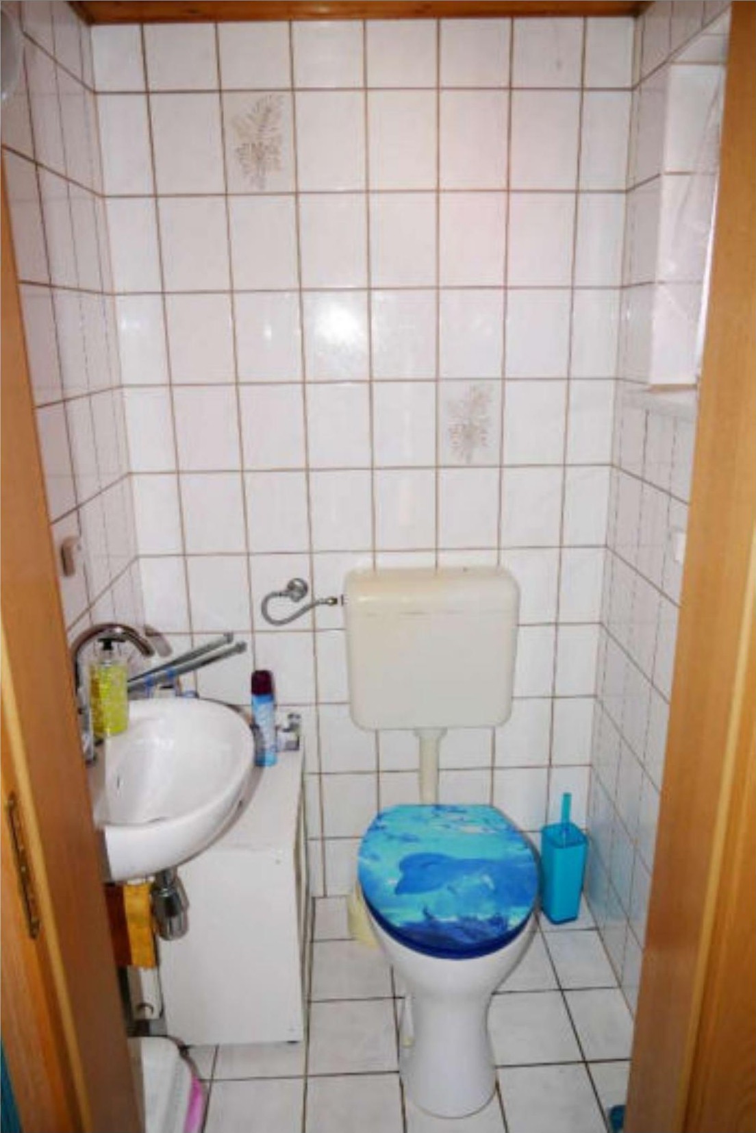 Badezimmer mit weiß gefliesten Wänden, Toilette mit blauem Sitz, Waschbecken und Toilettenbürste