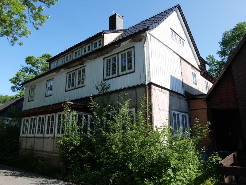 Seitenansicht eines zweistöckigen Hauses mit Holzverkleidung, mehreren Fenstern und Dachgauben, umgeben von Grünanlagen