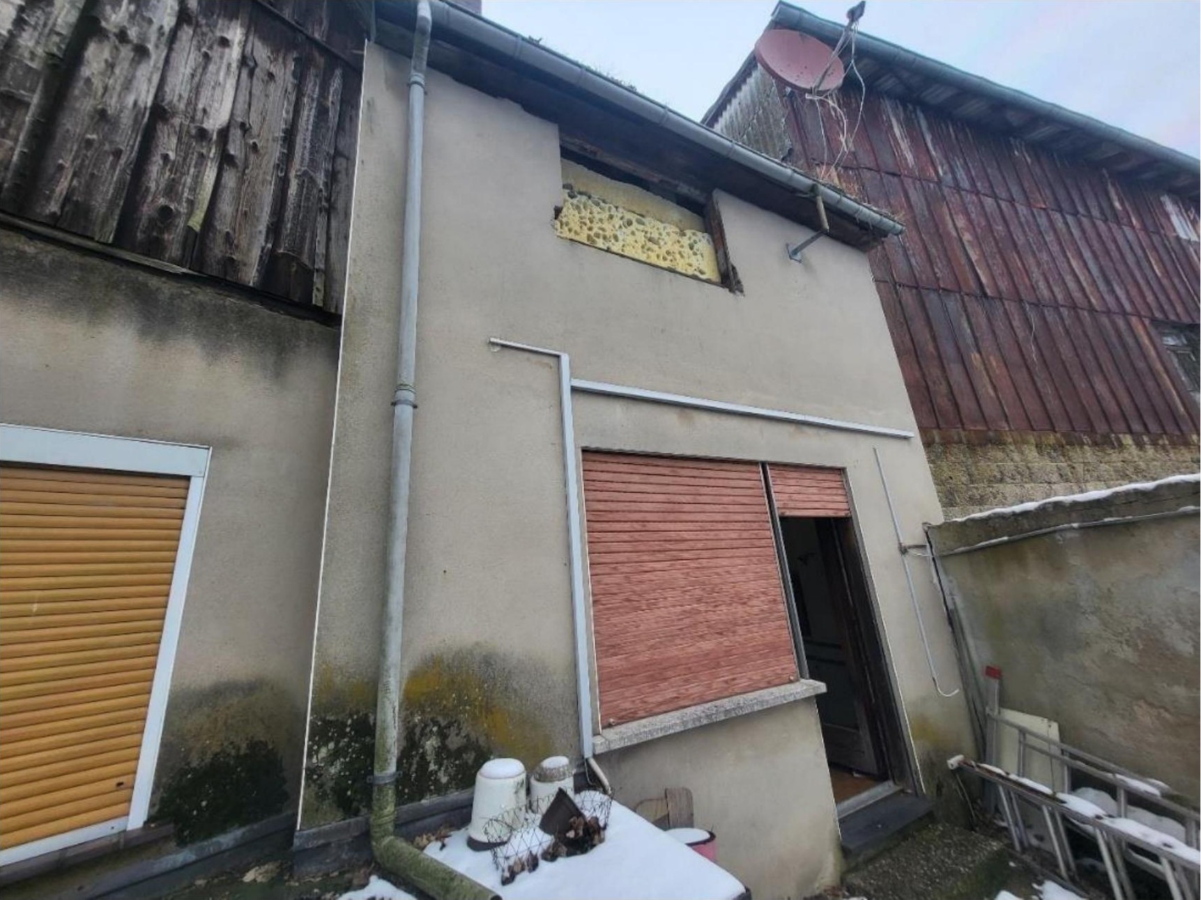 Rückansicht eines Gebäudes mit verwitterter Fassade, Rollläden, Tür und Schornsteinrohr; Schnee auf dem Boden und Dach