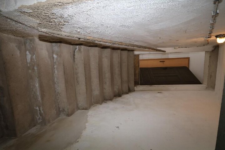 Kellerbereich mit Betontreppe, weiß gestrichener Decke und Boden, im Hintergrund eine dunkle Tür mit Holzrahmen