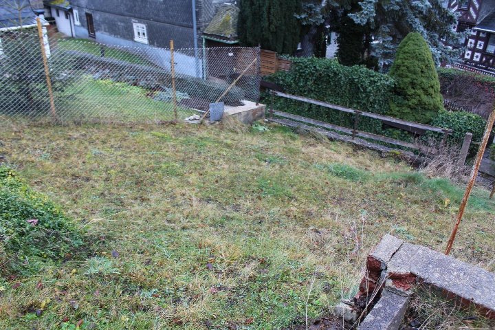 Gartenfläche mit Gras und vereinzelten Pflanzen, umgeben von Maschendrahtzaun und Holzgeländer, im Hintergrund Gebäude und Hecken in Meura, Ortsstraße 127