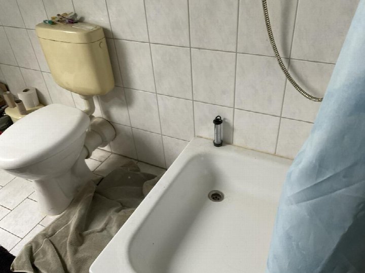 Badezimmer mit weißer Fliesenwand, WC mit cremefarbenem Spülkasten, Badewanne mit Duschschlauch und blauer Duschvorhang. Auf dem Boden liegt ein grauer Handtuch
