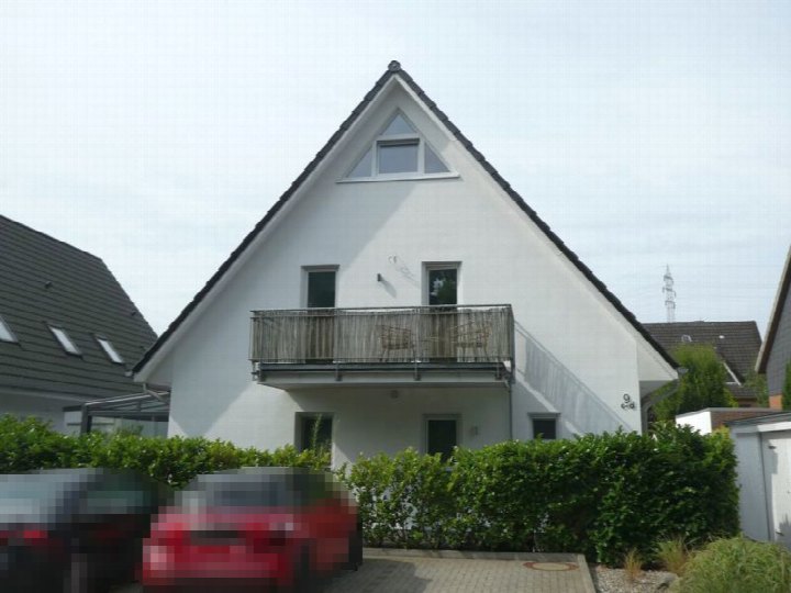 Fassade einer Eigentumswohnung in Scharbeutz, Kiepenberg 9, mit weißer Außenwand, schwarzem Satteldach, Balkon im Obergeschoss und grüner Hecke im Vorgarten